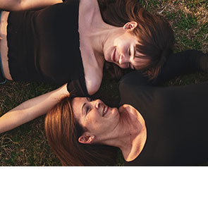 Une jeune femme (trentenaire) et une autre femme (cinquantenaire) brunes avec un t-shirt noire sont allongées dans l'herbe et se regardent en se souriant.