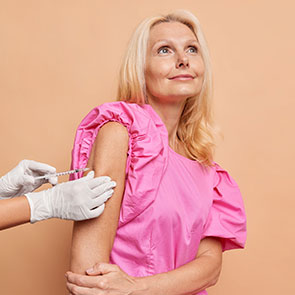 Une dame dans la cinquantaine/soixantaine portant un haut rose se fait vacciner.